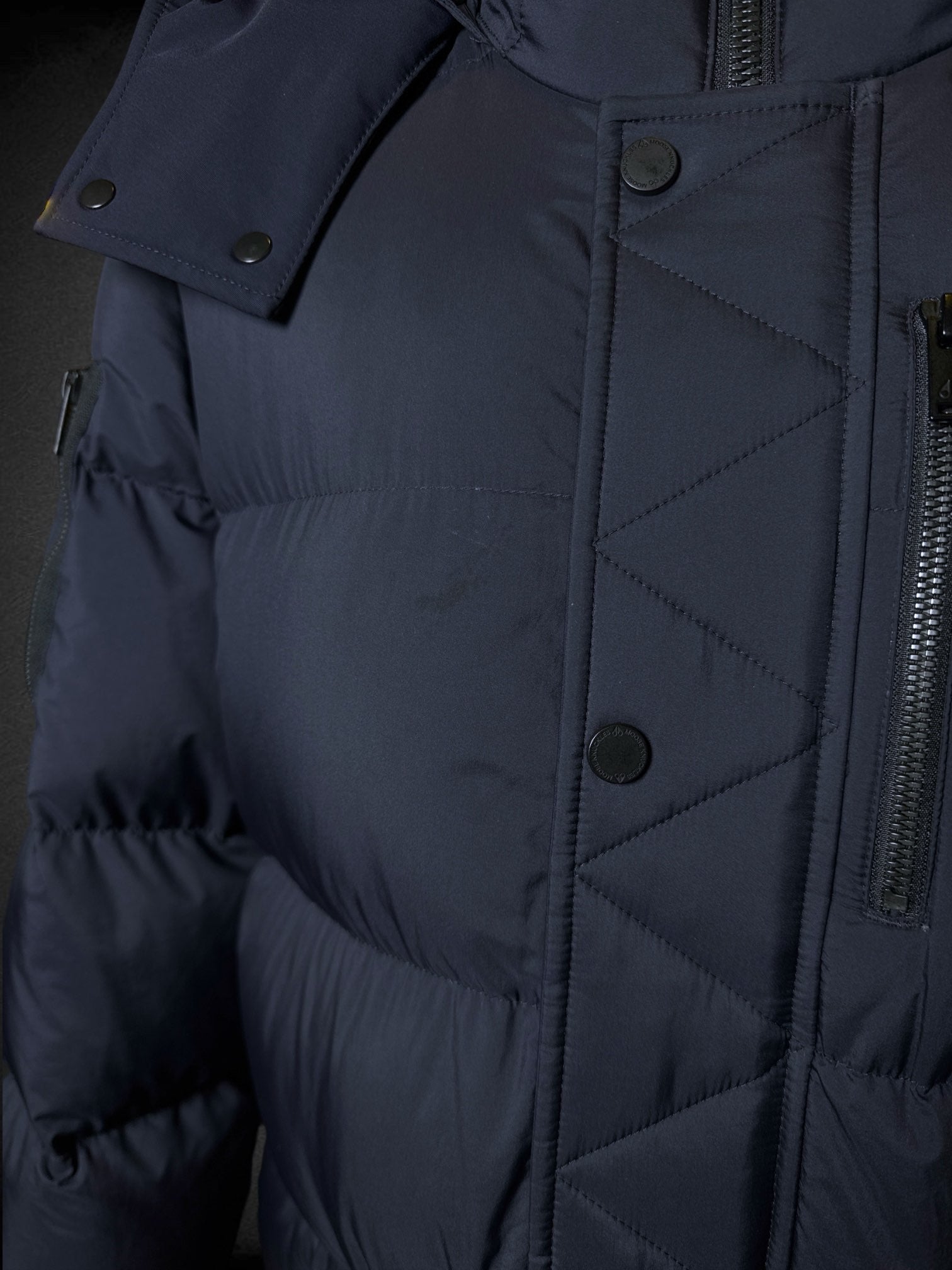 L Moose Knuckles dark blue puffer coat with zipper and snap button pockets, decorative slipper shaped like axe, and logo on sleeve